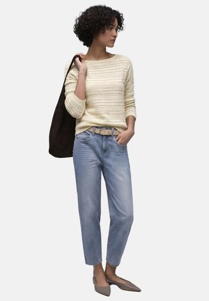 Vrouw met krullend haar, gekleed in een beige gebreide trui, lichtblauwe spijkerbroek, grijze platte schoenen, draagt een zwarte schoudertas, staand met haar hand in haar zak.