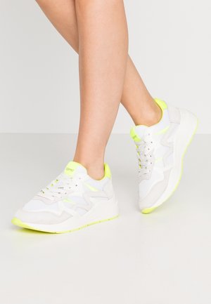 Beine mit weißen und neongelben Esprit-Sneakers mit Schnürsenkeln vor einem schlichten hellgrauen Hintergrund.