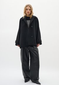 Black wool-blend oversized coat with double buttons, large lapels, and wrist accents, paired with high-waisted black leather trousers.
