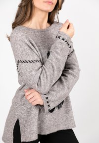 Grau gestrickter Pullover aus flauschigem Material mit schwarzem Stickmuster an den Ärmeln und einem hoch-niedrig geschnittenen Saum mit seitlichen Schlitzen.
