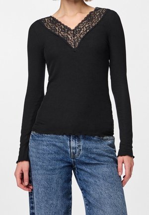 Femme portant un haut noir ajusté à manches longues avec un détail en dentelle en V au col et un jean bleu taille haute sur un fond uni.