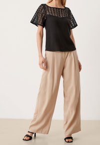 Haut en dentelle noire à manches courtes, corsage lisse, associé à un pantalon large beige et des sandales à talons noires. Fond neutre.