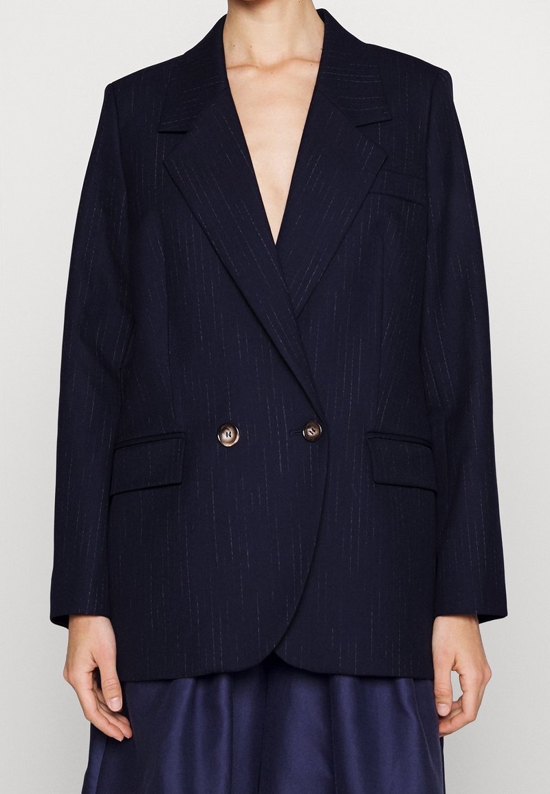 Femme portant un blazer croisé bleu marine foncé à fines rayures avec deux boutons et des poches à rabat, sur une jupe foncée.