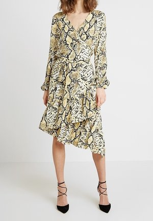 Femme portant une robe portefeuille jaune et noire à imprimé serpent, manches longues et ourlet asymétrique à volants, associée à des escarpins noirs à lacets, pointus.