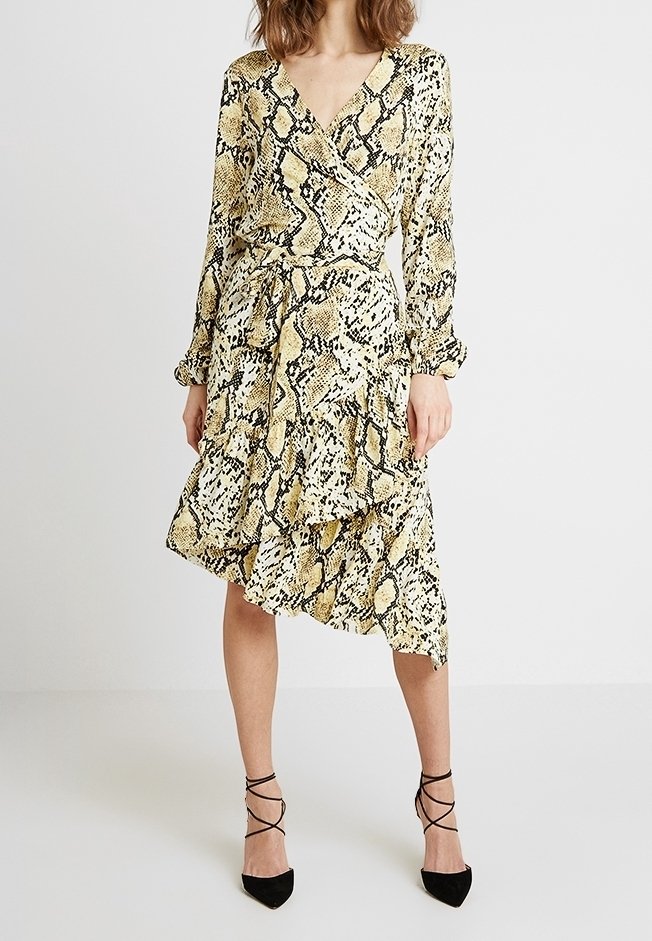 Femme portant une robe portefeuille jaune et noire à imprimé serpent, manches longues et ourlet asymétrique à volants, associée à des escarpins noirs à lacets, pointus.