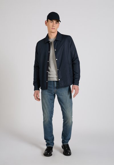 Hombre con gorra negra, chaqueta azul marino abotonada sobre suéter gris, vaqueros azules y zapatos negros, de pie contra un fondo liso.