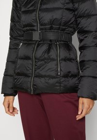 Czarna pikowana kurtka z paskiem i kieszeniami na zamek, noszona z ciemnoczerwonymi spodniami, ręce luźno opuszczone wzdłuż ciała na jasnym tle.