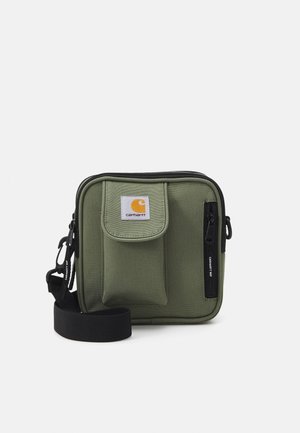 Sac d'épaule compact en tissu vert olive avec poche avant, poche zippée verticale, écusson avec logo Carhartt, et bandoulière noire réglable sur fond blanc.