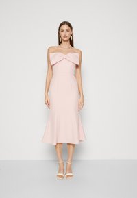 Abito midi in morbido tessuto rosa, design senza spalline con grande fiocco sul davanti, silhouette a gonna svasata, abbinato a tacchi bianchi con cinturino.