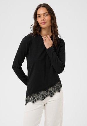 Femme portant un haut noir à manches longues avec une bordure en dentelle à l’ourlet et un pantalon blanc, debout devant un fond clair et uni.