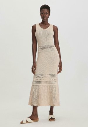 Ärmelloses Strickkleid in Beige mit Durchbrüchen, einem taillierten Oberteil, ausgestelltem Saum und gestuften Spitze-Details. Kombiniert mit hellen Sandalen.