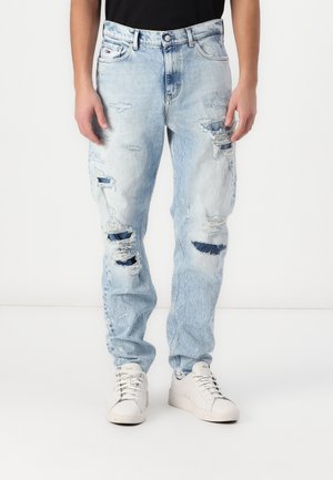 Jasnoniebieskie podarte jeansy z wieloma rozdarciami, noszone z białymi trampkami i czarną bluzką, pokazane od pasa do stóp.