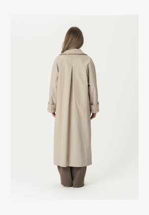 Beige pikas jope kraega, millel on volditud tagaosa ja rullitud varrukad. Valmistatud siledast kangast, kantud pruuni pükstega.