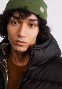 Giacca a vento nera con colletto alto, maglione a maglia arancione-marrone sotto, e berretto a maglia verde con un logo. Capelli ricci e piercing sul viso.