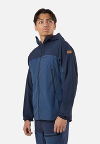 Jonge man draagt een tweekleurig blauw zip-up hoodie-jas met ritssluitingen aan de zakken, kijkt iets naar de zijkant, staande tegen een effen achtergrond.
