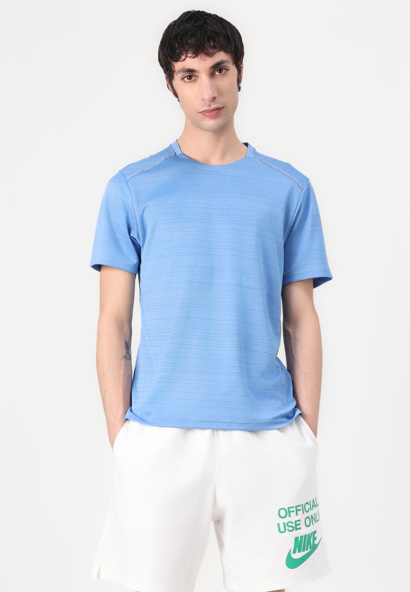 Camiseta deportiva de manga corta en azul claro con un patrón texturizado y cuello redondo, combinada con pantalones cortos blancos que presentan una marca verde.