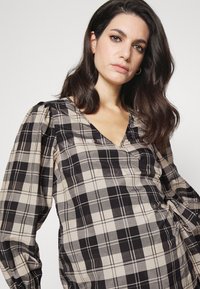 Blusa xadrez preta e bege com decote em V e mangas bufantes, feita de um tecido suave com um brilho sutil e um detalhe na cintura ajustável.
