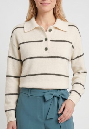 Femeie purtând un pulover crem cu dungi orizontale închise la culoare și trei nasturi, asortat cu pantaloni teal cu talie înaltă legați cu o curea în formă de fundă.