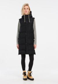Schwarze Steppweste mit Kapuze, ausgestattet mit Druckknöpfen, zwei Fronttaschen und kombiniert mit einem grauen Langarmshirt und schwarzen Leggings.