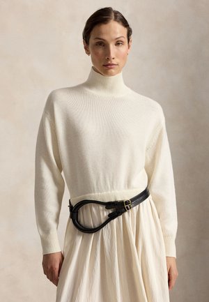 Femme portant un pull à col roulé crème, une longue jupe crème plissée, une ceinture noire et des bottines en daim beige, debout sur un fond neutre.