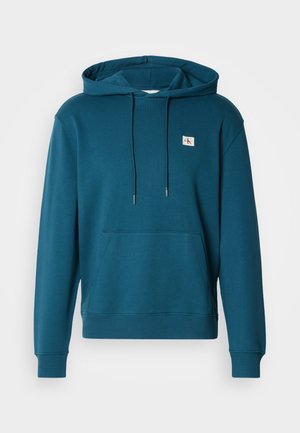 Teal hettegenser laget av mykt materiale, med en frontlomme og en liten logopatch på brystet. Snorene er festet til hetten.