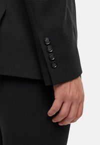 Ärmel einer schwarzen texturierten Jacke mit vier schwarzen Knöpfen mit floralen Gravuren. Nahaufnahme, die eine Hand zeigt, die die Jacke hält.
