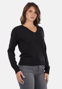 Maglione nero a V realizzato in un tessuto morbido con polsini e orlo a coste. Presenta un design aderente e maniche lunghe. Indossato con jeans grigi.
