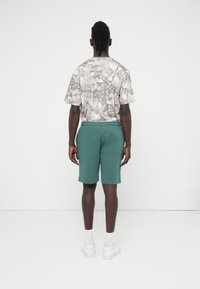 Κοντομάνικο, γκρι μπλουζάκι με σχέδια· πράσινο σορτς με ελαστική μέση· εμφανίζονται επίσης άσπρα αθλητικά παπούτσια και κάλτσες. Πλάγια όψη από πίσω.