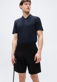 Σκούρο μπλε πόλο με φερμουάρ στο γιακά και λεπτομέρεια λογότυπου, σε συνδυασμό με μαύρα σορτς. Το ύφασμα φαίνεται λείο και ελαφρύ. Κράτα γκολφ στο χέρι.