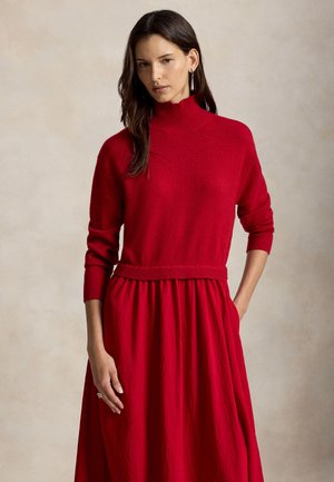 Femme aux longs cheveux foncés portant un pull à col roulé rouge et une jupe rouge assortie sur un fond beige uni.