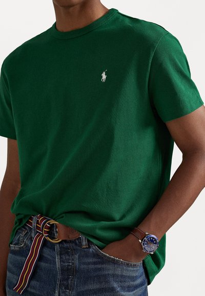 Camiseta de algodón verde con cuello redondo y un pequeño logo blanco en el lado izquierdo del pecho, combinada con jeans de mezclilla azules y un cinturón multirayas.