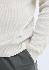 Pullover bej cu tricotaj, cu manșete și tiv cu nervuri, evidențiind un material texturat. Include un label mic cu text care detaliază filosofia brandului.