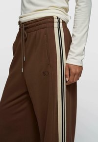 Joggers marron avec des bandes latérales noires et crème, taille à cordon, et détail brodé "10" sur le devant. Texture de tissu douce.