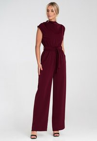 Burgundowy kombinezon z krótkimi rękawami, wysokim kołnierzem i paskiem w talii. Wykonany z gładkiego materiału, z szerokimi nogawkami i bez widocznych wzorów.
