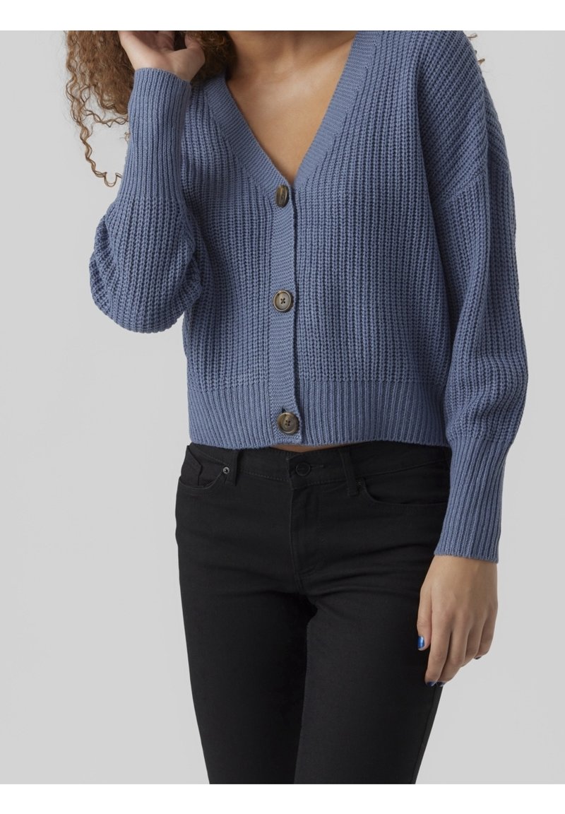 Blå stickad cardigan med v-ringning, tre knappar och ribbad textur, matchat med svarta byxor. Cardiganen har vida ärmar och en kortad passform.