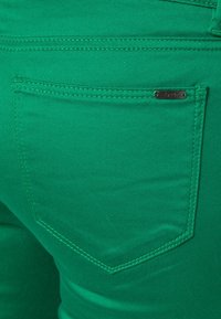 Groene denim jeans met een achterzak met een metalen label. De stof heeft een gladde textuur en de stiksel is uniform en in dezelfde kleur.