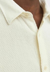 Chemise à manches courtes de couleur crème avec un motif texturé en nid d'abeille et un col. Comprend une patte de boutonnage.