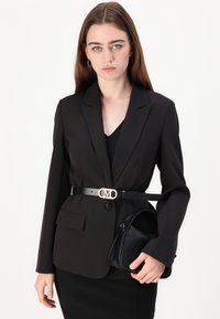 Svart blazer med revers, tettsittende design og to frontlommer. Båret over en svart topp, komplementert av et bredt belte og en svart veske.