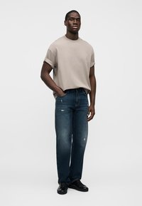 Μπεζ T-shirt με ρολαρισμένα μανίκια και σκούρο μπλε τζιν, με ελαφρύ φθαρμένο εφέ. Το μοντέλο φοράει μαύρα παπούτσια και στέκεται μπροστά σε μονόχρωμο φόντο.