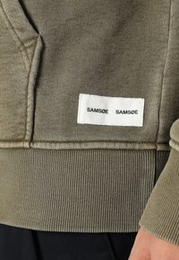 Olivgrüner Sweatshirt mit gerippten Bündchen und Saum, versehen mit einem weißen Stoffetikett mit dem Text "SAMSOE SAMSOE". Weiche Textur und lässiges Design.