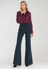 Kobieta ubrana w fioletową bluzkę z falbanami, wpuszczoną w czarne spodnie z wysokim stanem o szerokich nogawkach oraz czarne szpilki, pozująca na jednolitym tle.