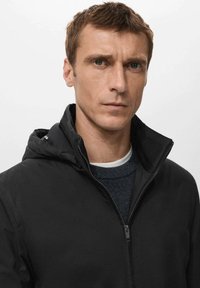 Giacca nera con cappuccio e zip frontale, dotata di colletto alto e tessuto leggero e liscio. Indossata sopra un maglione scuro.