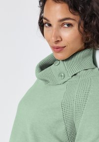 Mátově zelený svetr s vysokým límcem, který má texturovaný pletený vzor a dva knoflíky na boku. Měkká látka s žebrovanými akcenty.