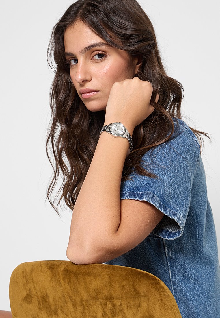 Junge Frau mit langen dunklen Haaren, die eine silberne Armbanduhr und ein blaues Denim-Hemd trägt, während sie ihren Arm auf einem senfgelben Stuhl ruht.