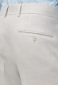 Pantalon de costume gris clair avec une poche arrière boutonnée et des passants pour ceinture, porté avec une chemise bleu clair.