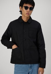 Svart knappjakke med krage, frontlommer og en glatt tekstur. Brukt sammen med blå denim jeans.