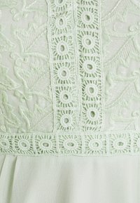 Světlý zelený textil s detailním květinovým vyšíváním a čtvercovými krajkovými vzory, který obsahuje listové motivy a kruhové otvory v krajkovém lemování.