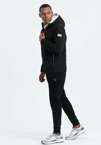Veste softshell noire avec une capuche grise, poches zippées et pantalon de survêtement noir. Baskets grises et blanches avec des accents en maille.