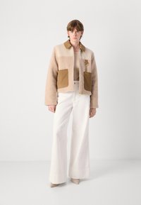 Veste en fausse fourrure beige et marron clair, coupée, avec des poches en velours côtelé, une fermeture éclair dorée et un col. Associée à un pantalon blanc évasé.