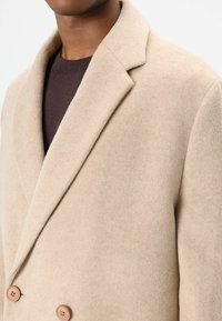 Beige double-breasted jas van zachte stof, met een notched kraag en lichtbruine knopen, getoond op een donker trui.